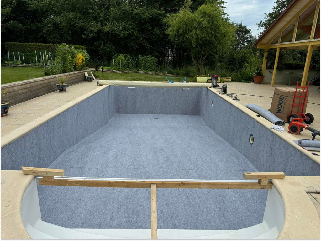 Piscine en construction avec feutrine grise installé, terrasse en pierre et équipements de chantier visibles