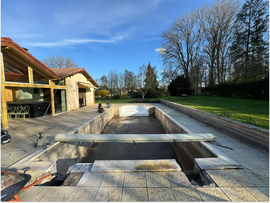 Piscine vide en béton brut avec margelles posées, maison contemporaine et jardin en arrière-plan