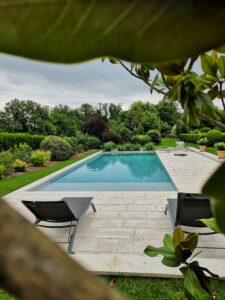 Piscine rectangulaire moderne avec terrasse en pierre naturelle, chaises longues noires et jardin paysager avec pelouse et massifs arbustifs